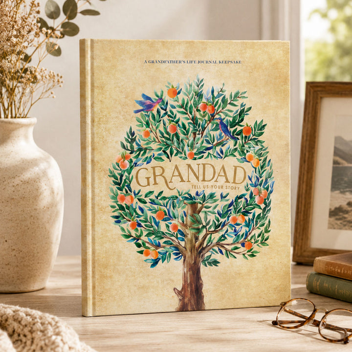 A hardcover life keepsake journal titled 'Grandad Tell Us Your Story' stands upright on a wooden table. The cover features a vintage-style illustration of a tree with orange fruits and blue birds on a warm beige background.