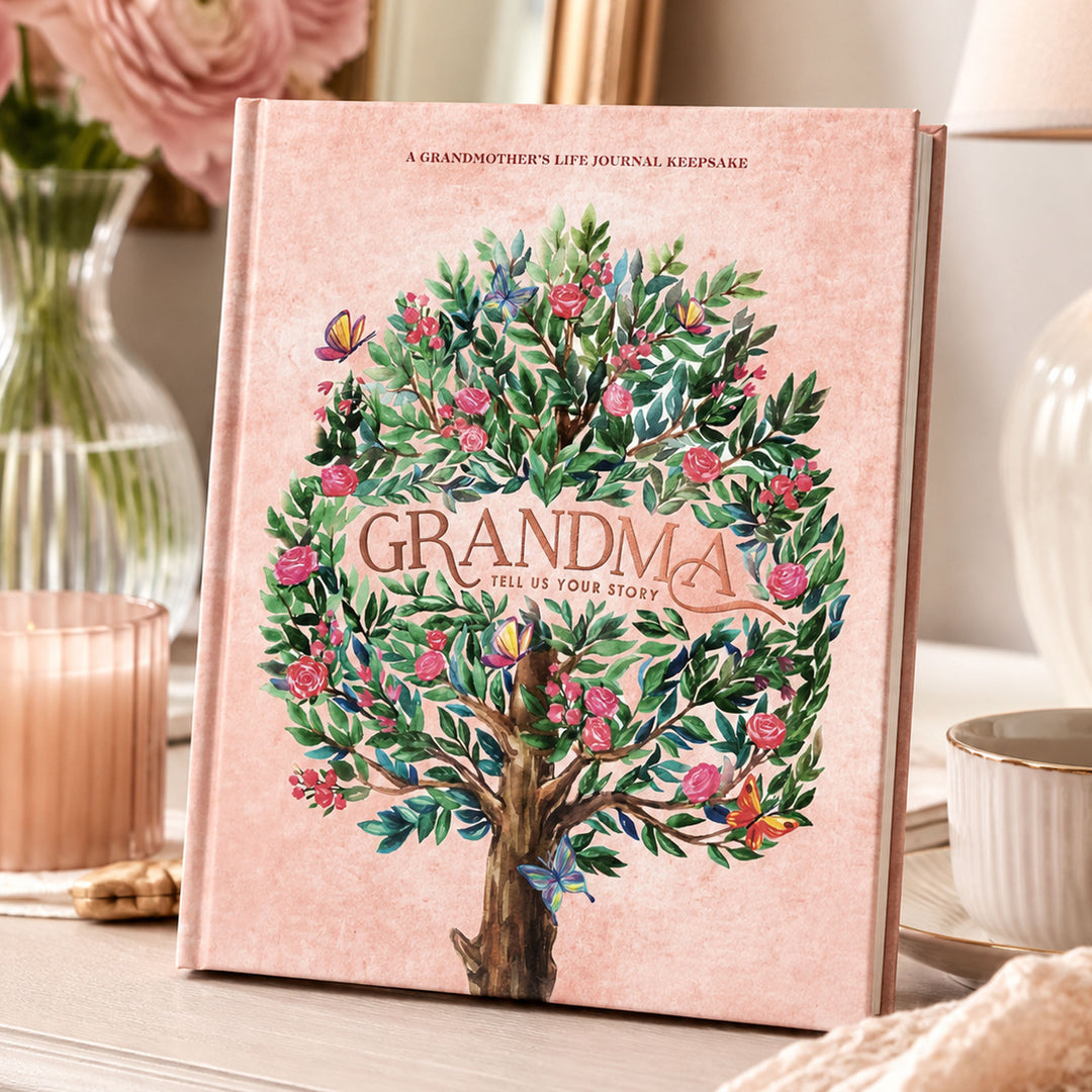 A pink hardcover life journal titled 'GRANDMA: TELL US YOUR STORY' featuring a detailed illustration of a tree with pink flowers and green leaves, styled on a tabletop with home decor props.